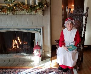 Mrs. Claus seated next to a fire in the Manor House fireplace, holding a book on her lap